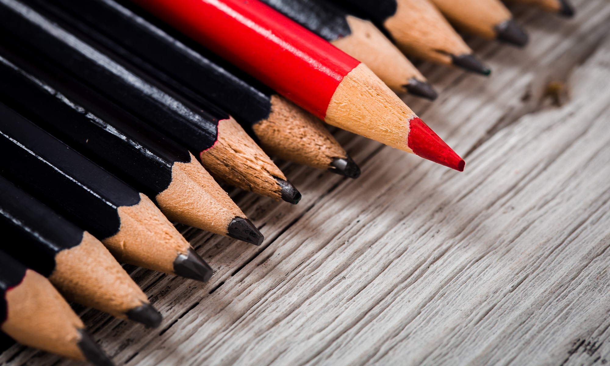 Roter Bleistift zwischen mehreren schwarzen Bleistiften auf einem Holztisch, der sich deutlich von der Gruppe abhebt und Individualität oder Leadership symbolisiert